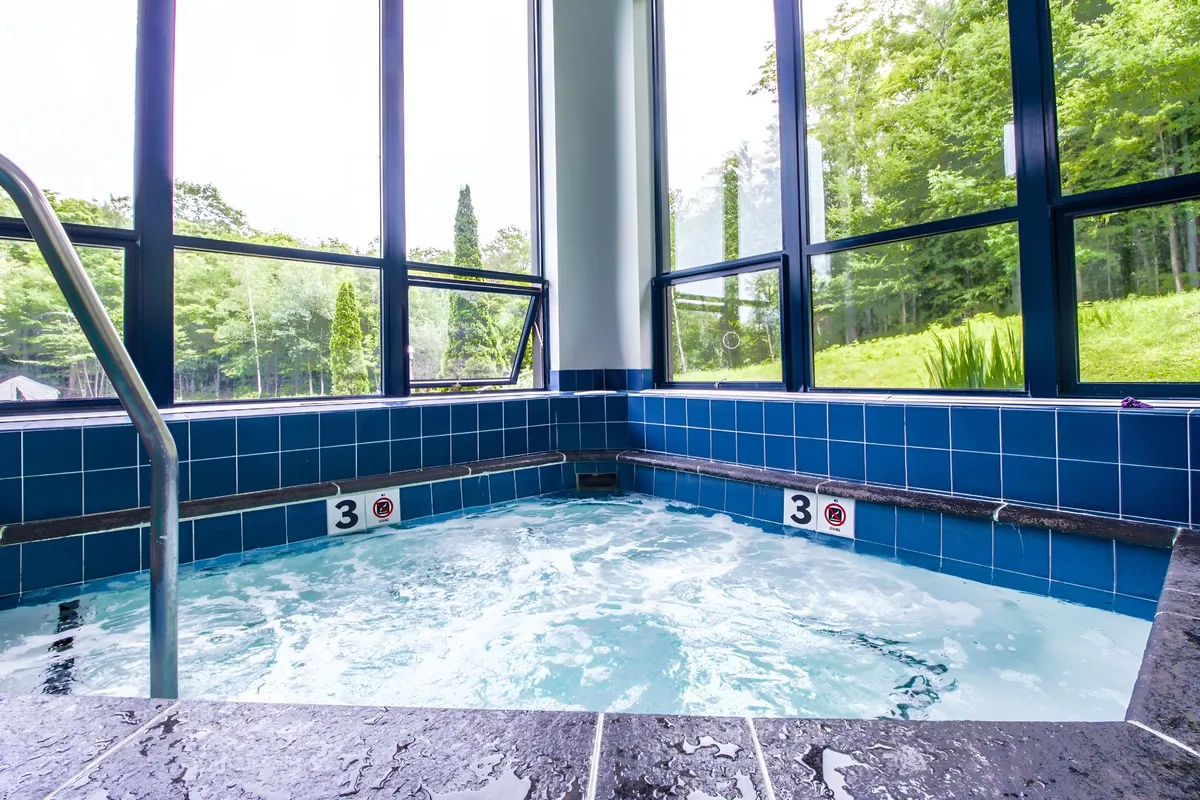 A tranquil hot tub room with oversized windows offers views of lush greenery outside. The setting feels calm and social, ideal for unwinding in the warmth while staying connected to the beauty of the outdoors.