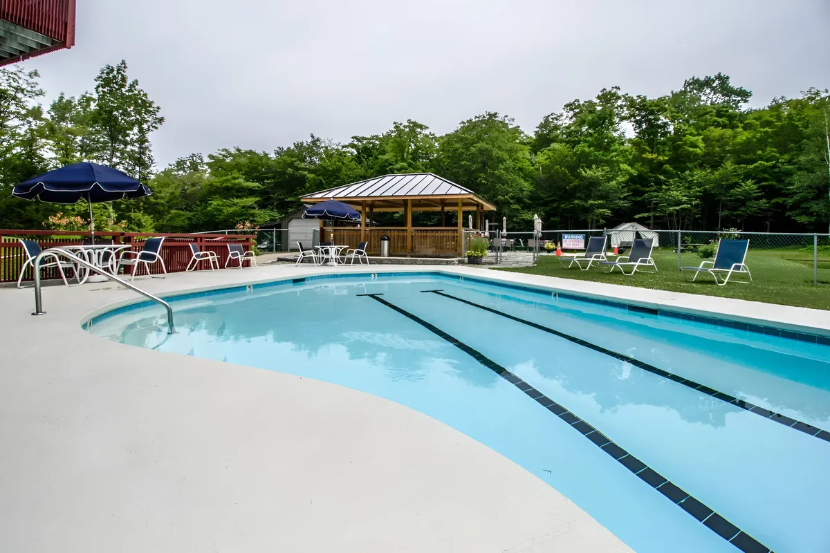 A vibrant outdoor pool scene featuring a sparkling swimming pool surrounded by lounge chairs, a gazebo, umbrellas, and lush landscaping. The setting suggests relaxation and wellness options available in the warmer months.
