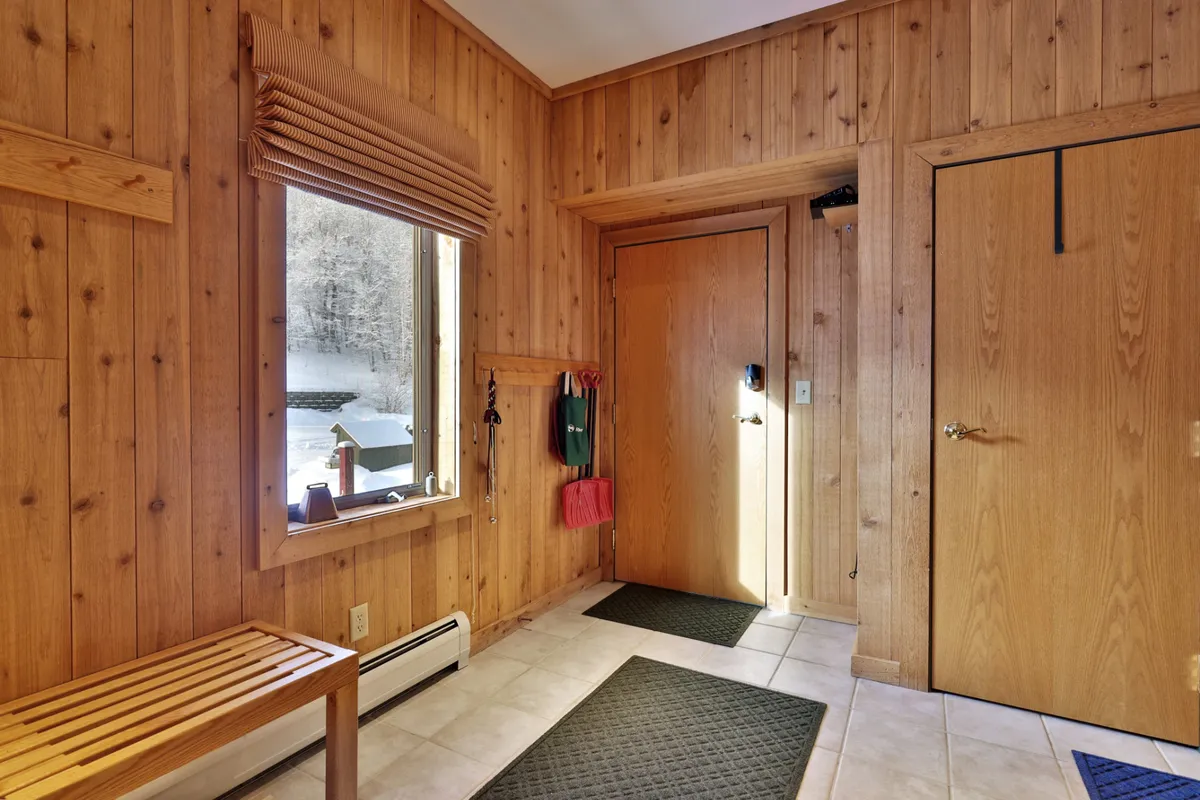 A cozy entry area lined with inviting wood paneling, a well-placed bench for gearing up or removing boots, and a window that frames the snowy outdoors. Floor mats and practical shelving make this the perfect staging area for outdoor adventures.