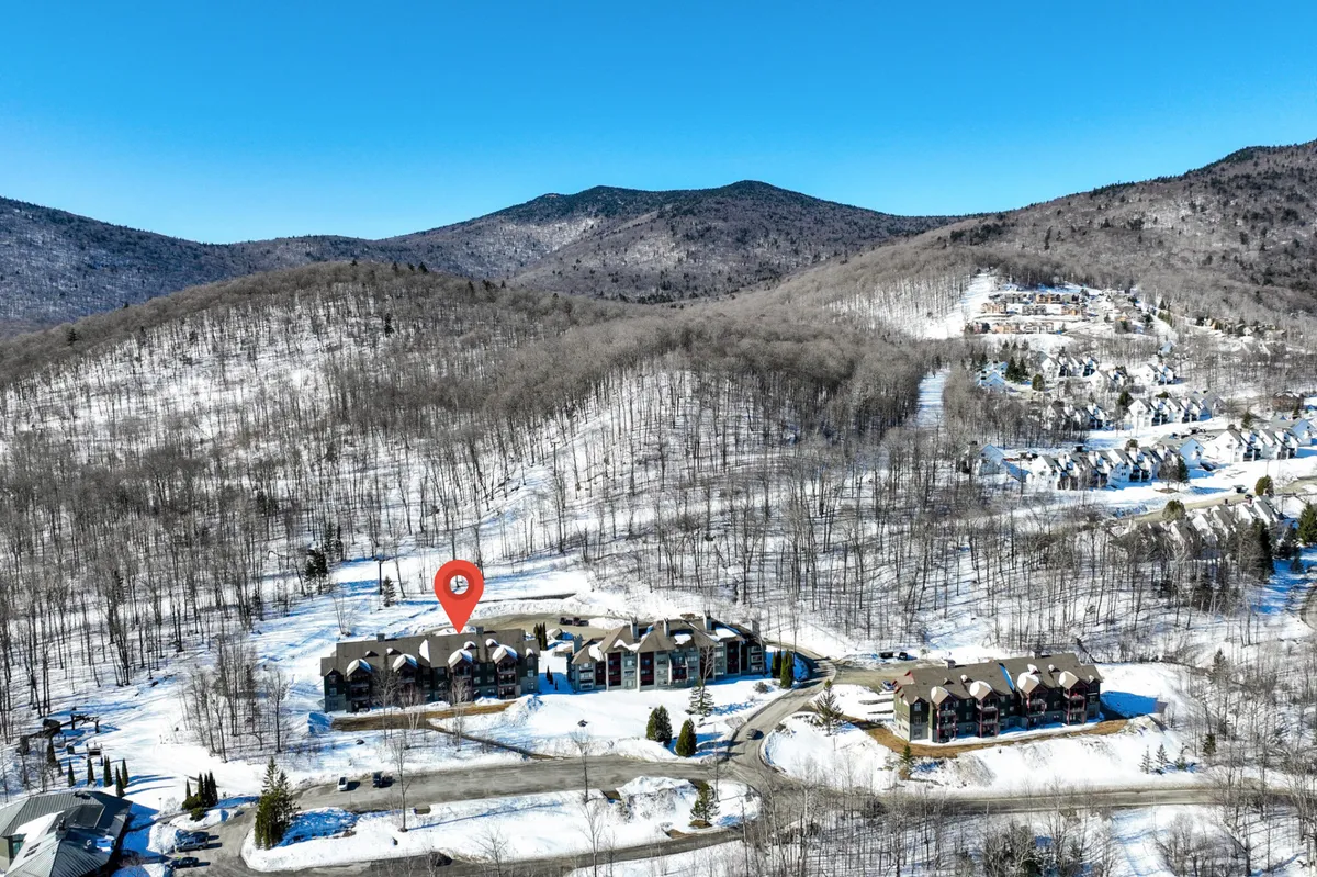 A wide exterior view of residential buildings nestled at the base of snow-covered mountains, with pine trees and winding roadways framing the winter scene. The landscape suggests year-round access to hiking and skiing trails from the doorstep.