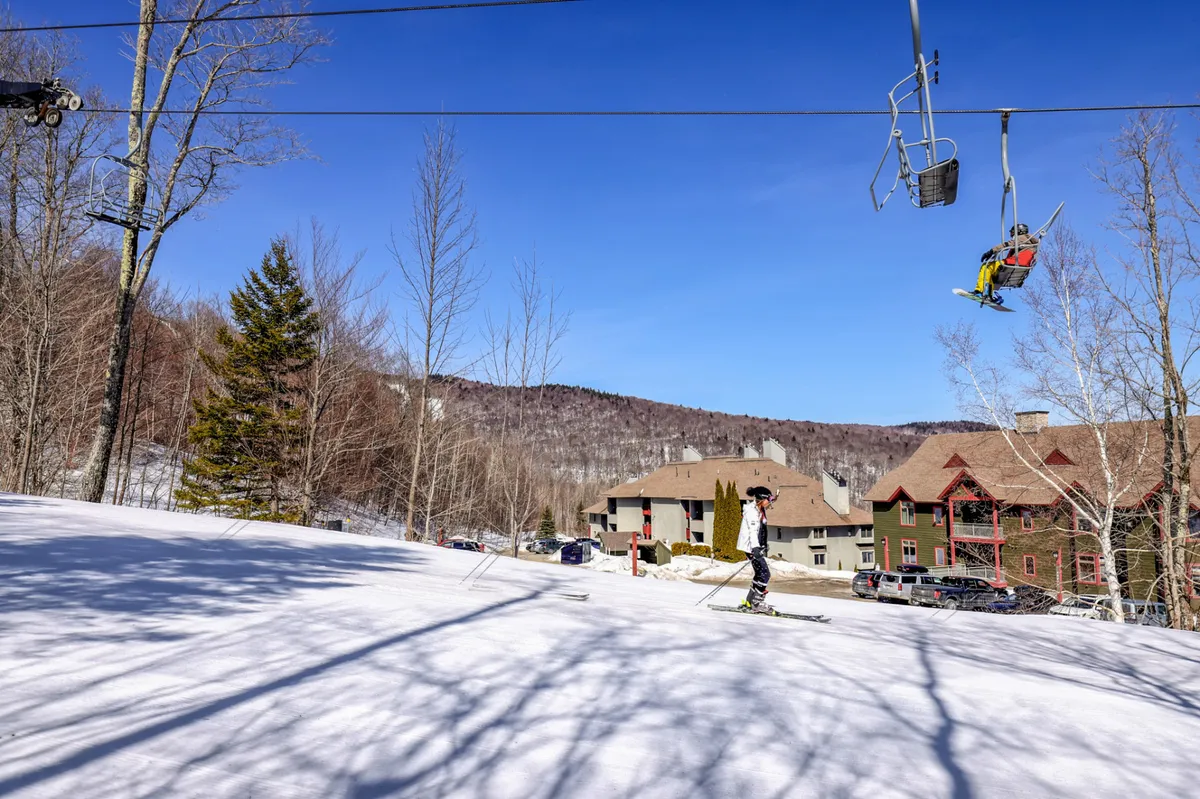 This dynamic exterior shot captures a ski lift in motion, a skier ready to descend, and the striking mountain backdrop of the resort. Surrounding buildings evoke the lively, adventurous spirit of a ski destination, while the mountains hint at year-round activities on the trails.