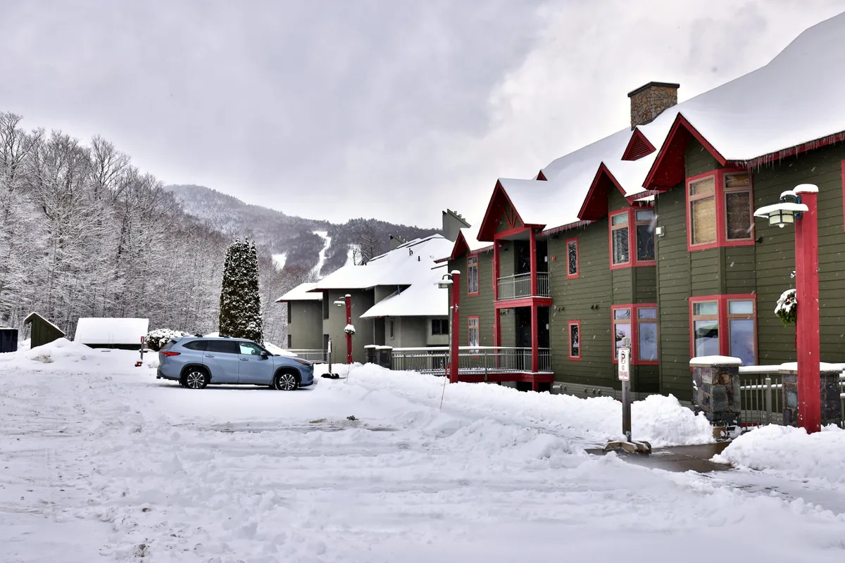 A wintertime exterior view of the condo complex, with snow-covered buildings, a frozen landscape, parked cars, and an evergreen tree. The scene is tranquil yet lively, hinting at nearby ski amenities and après-ski adventures.