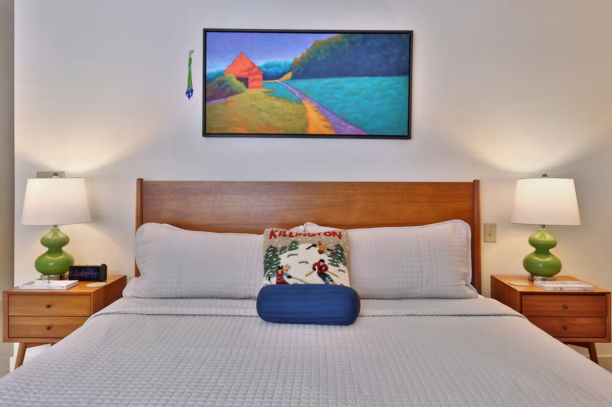 A bright bedroom featuring a plush bed flanked by nightstands and lamps, with modern artwork above the headboard. Sunlight streams in, creating a calm and inviting atmosphere ideal for unwinding after a day outdoors.