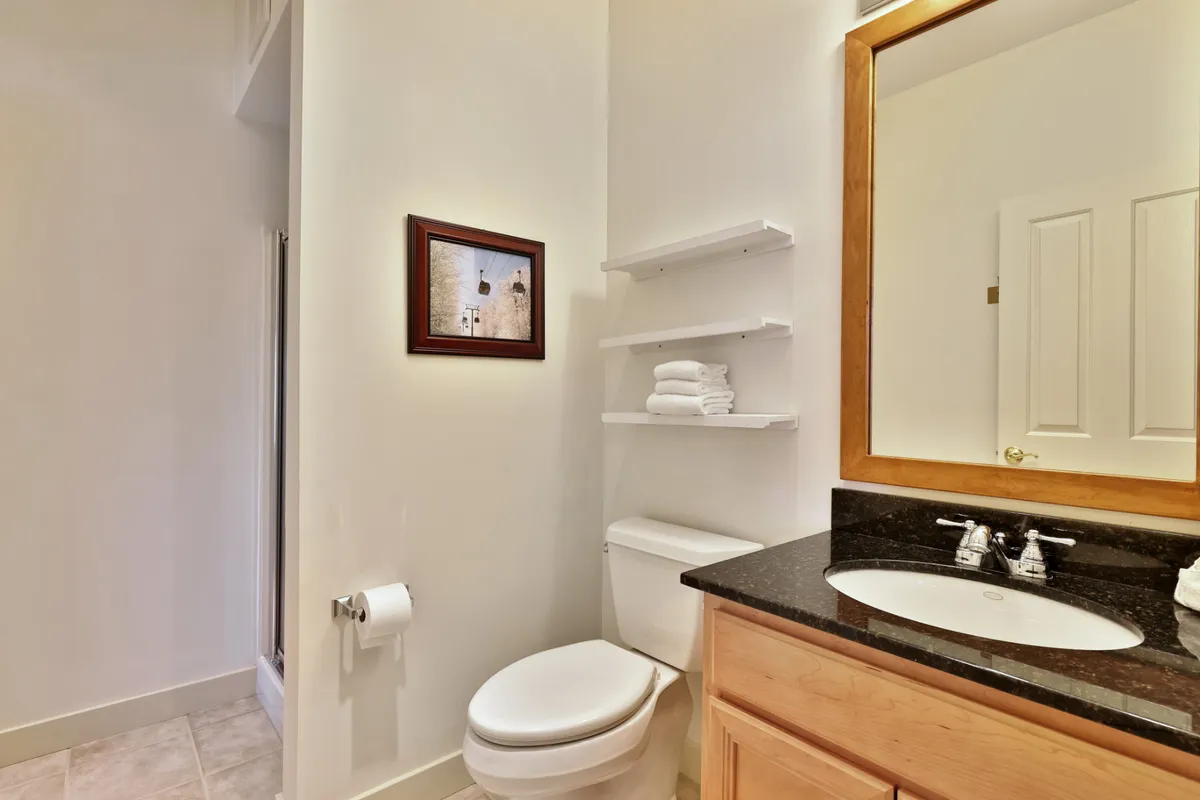 A neat bathroom with a toilet, sink, mirror, framed art, and open shelves neatly stacked with fresh towels. The design is functional yet calming, suited for guest comfort and convenience.