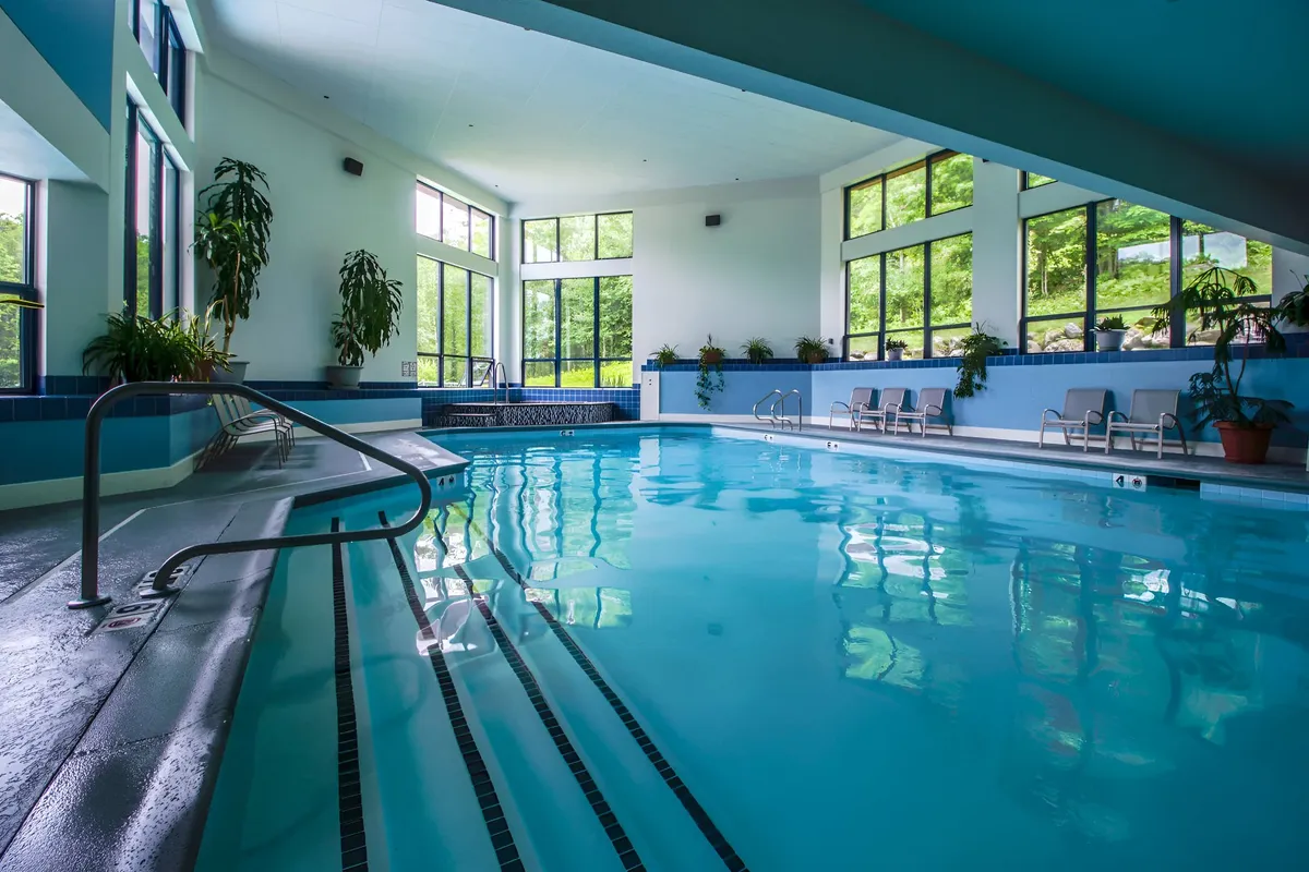 A spacious indoor swimming pool area featuring large windows that let in plenty of natural light, comfortable seating along the poolside, and lush green plants that accentuate the tranquil ambiance. The pool is the centerpiece, inviting guests to swim or simply relax by the water.