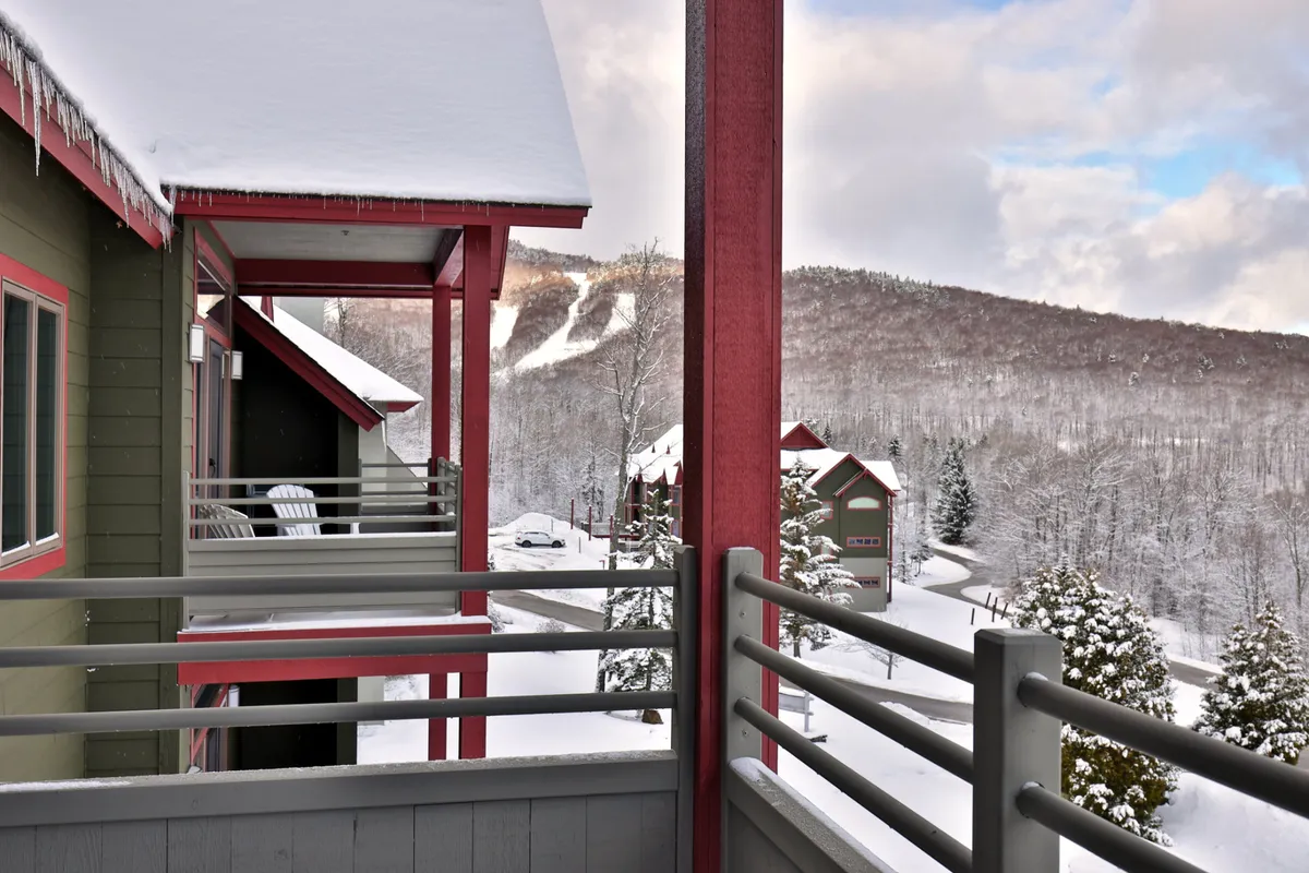 A sweeping view of snow-covered rooftops, distant ski slopes, wooden railings, and pine trees. The mood is serene, inviting groups to relax after a day of adventures or to start the morning with coffee overlooking the mountains.