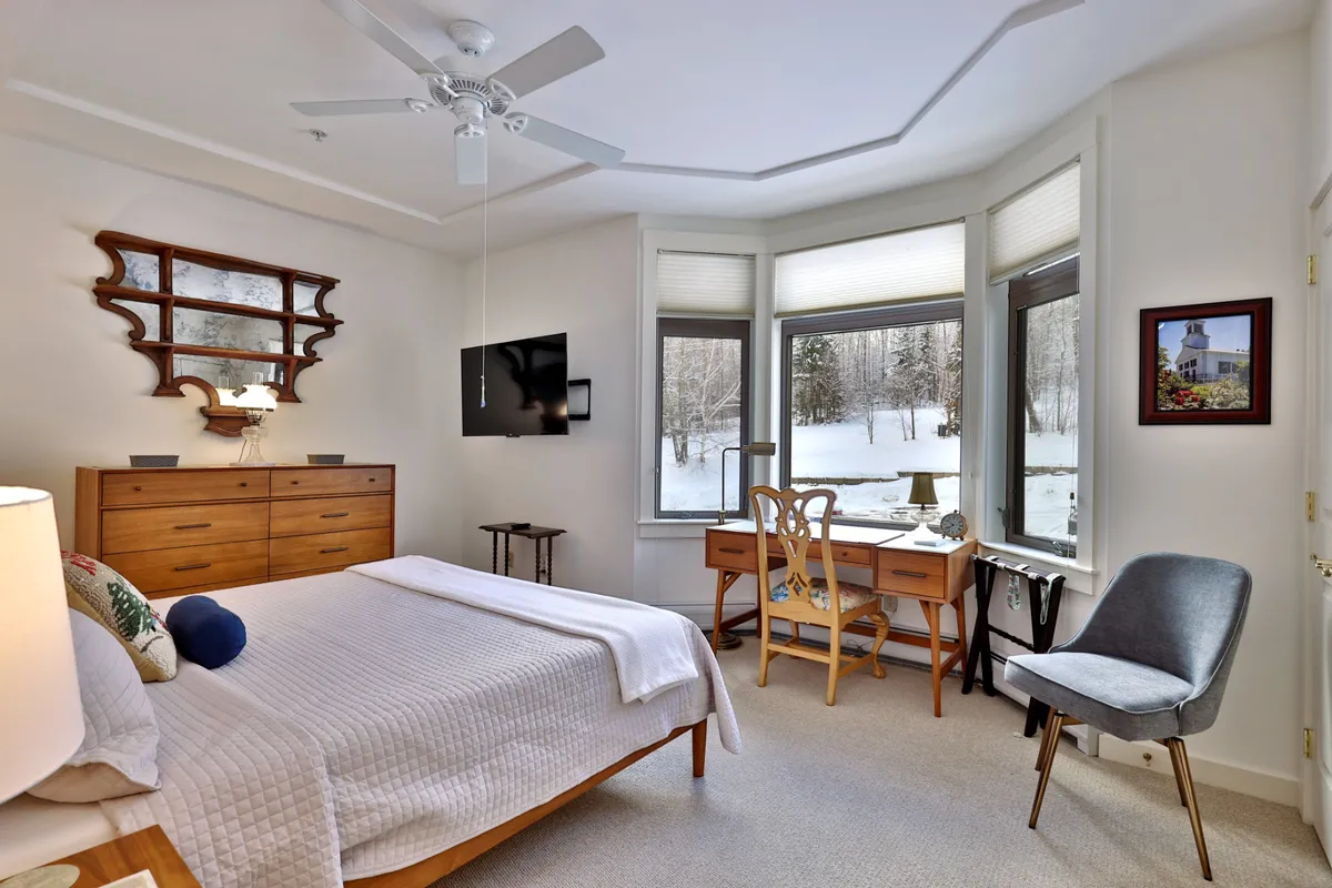 A tranquil bedroom scene with a cozy bed, a writing desk positioned by the window, a decorative mirror, and a dresser. The window offers peaceful views, adding to the room’s restorative character.