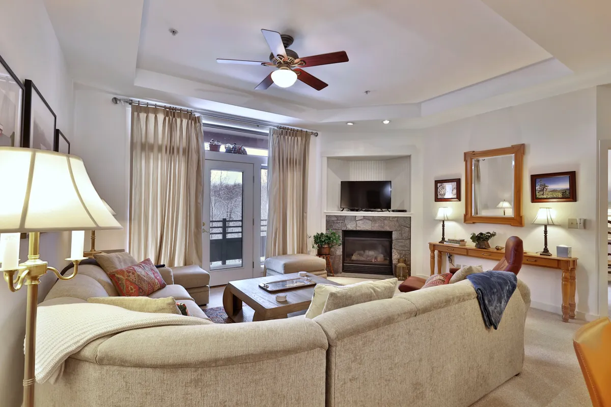 A spacious, cozy living area featuring plush sofas, a roaring fireplace, a large television, and expansive windows that let in natural light and offer snowy views. Decorative curtains add a touch of style, making the room perfect for relaxation or gathering after a day outdoors.