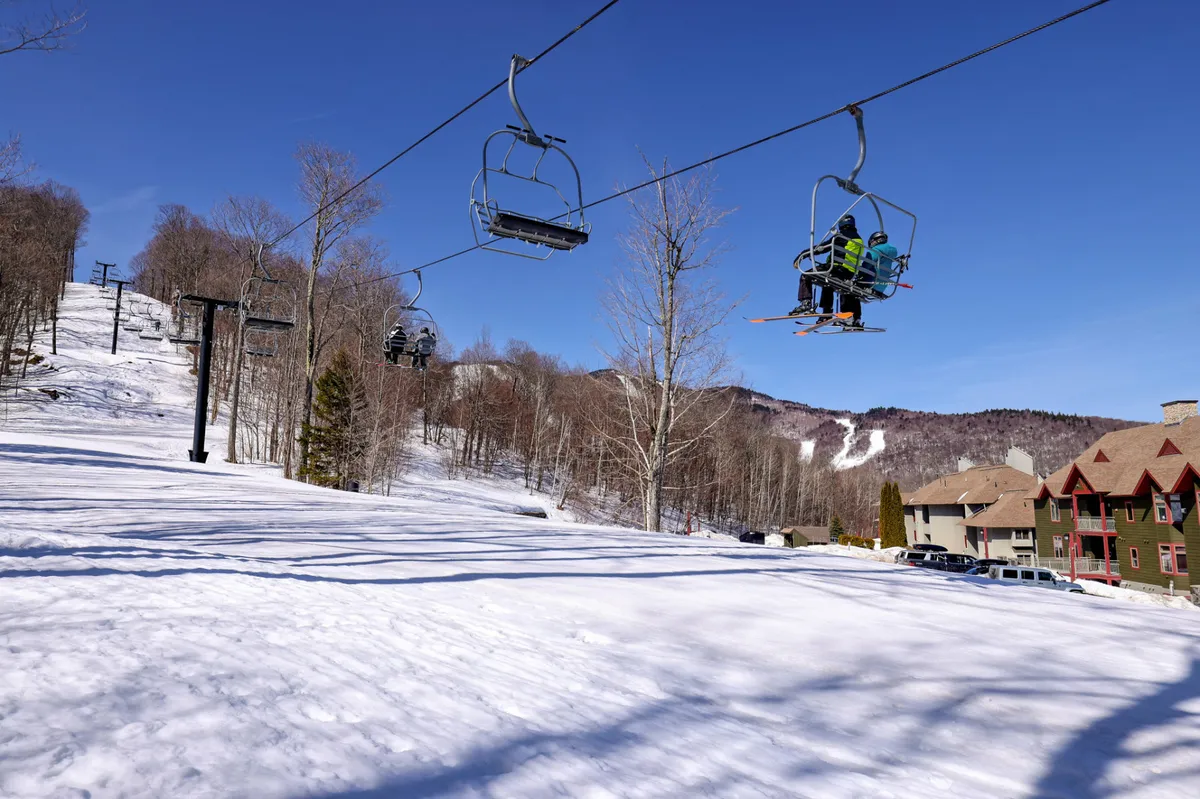 A vibrant exterior scene showing a ski lift ascending a snow-covered slope, surrounded by trees and holiday homes. Skiers and snowboarders are visible, enjoying the immediate access to the slopes. The area exudes both excitement and tranquility, with cozy lodges nestled at the base of the mountain.