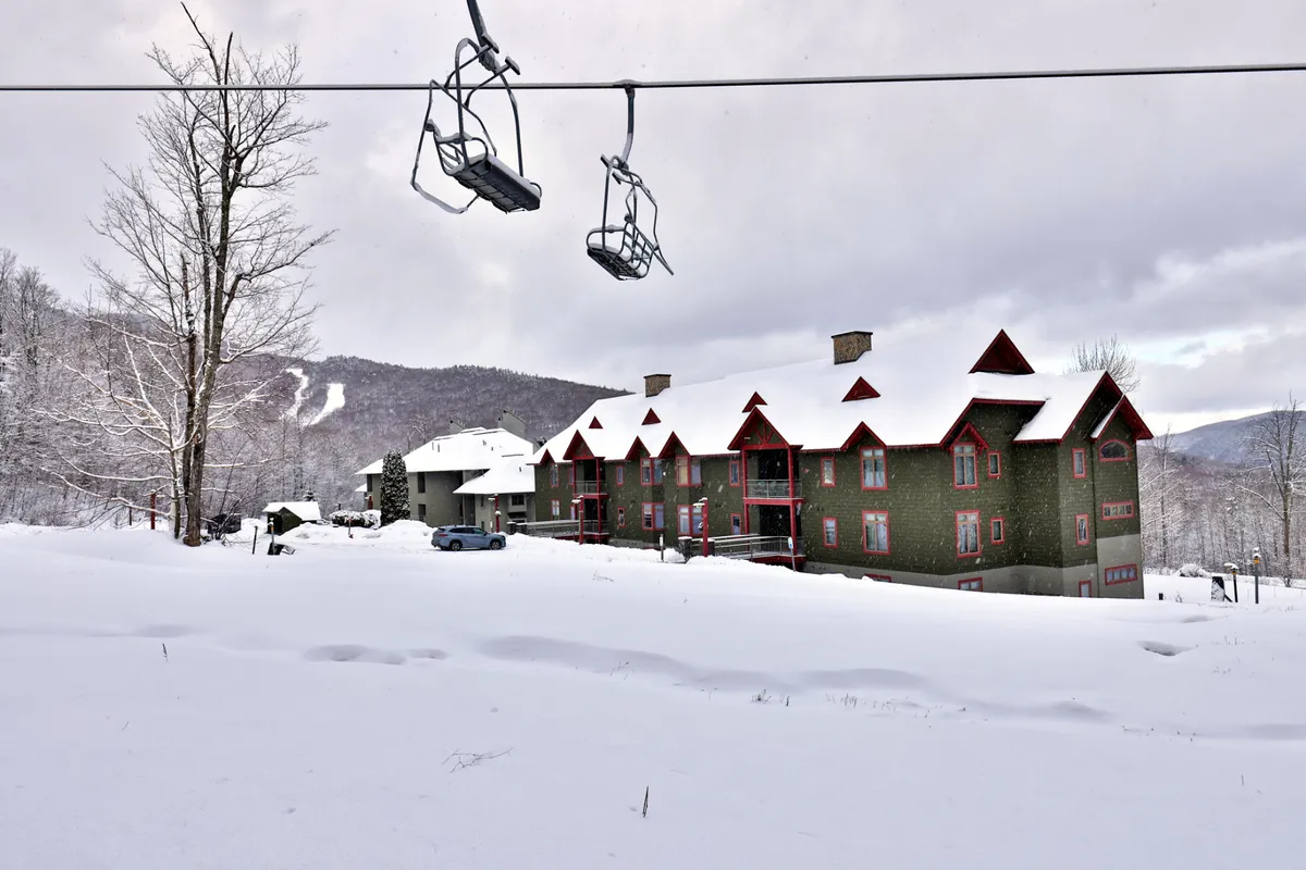A serene exterior shot highlighting the lodge with red accents, a snow-covered landscape, and a ski lift overhead. The composition captures the inviting base of the ski area with easy trail access and a welcoming atmosphere for groups arriving or returning from the slopes.