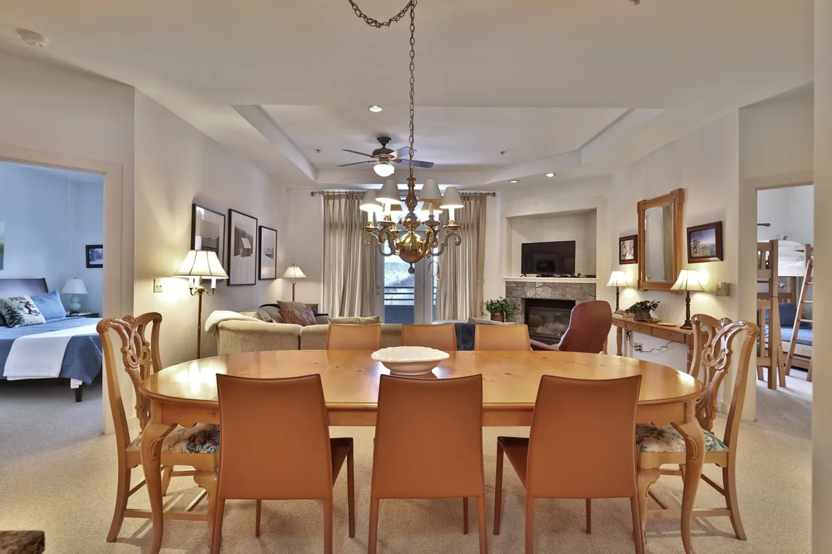 A wide view of the dining area with a large table set beneath a chandelier, comfortable sofas grouped near a fireplace, and bunk beds visible in the background. The setup welcomes family dinners, lively games, and cozy fireside chats, glowing in warm, inviting light.