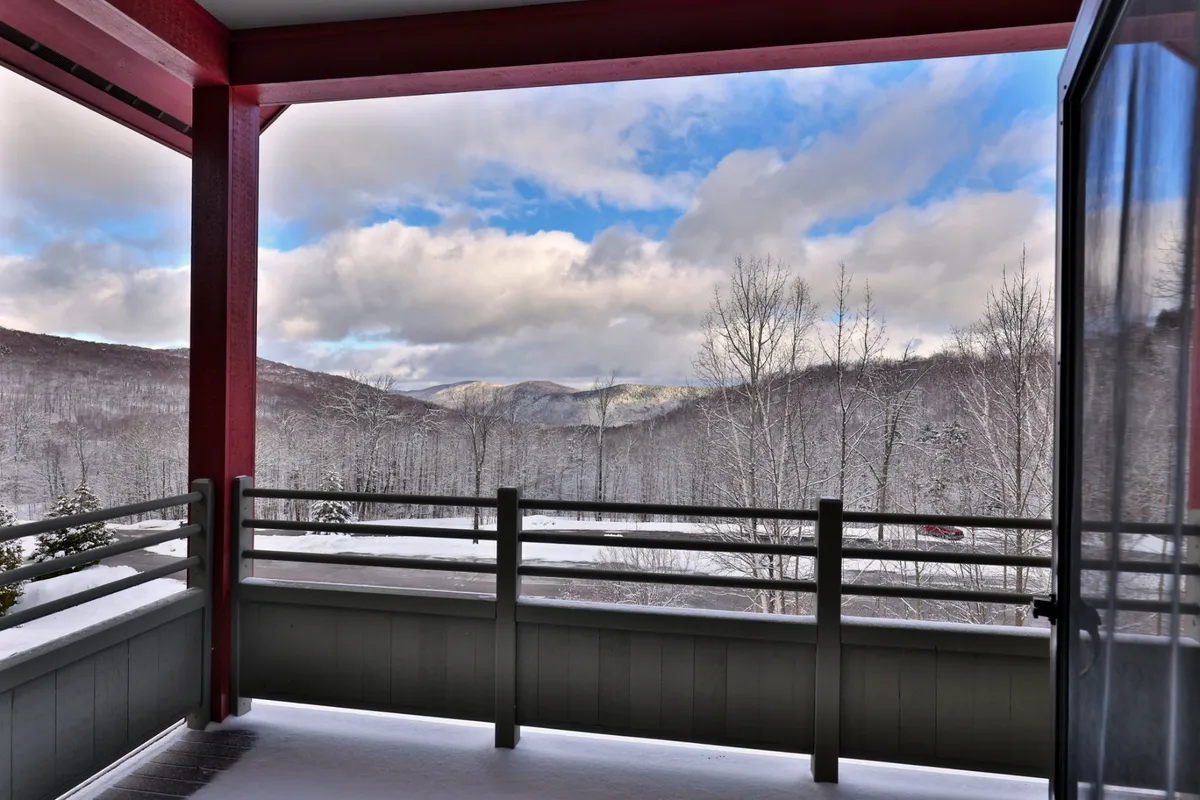 A balcony view overlooking snow-capped mountains, bare winter trees, and a blue sky, with a red railing in the foreground. The view suggests a tranquil spot to relax with coffee while soaking in the scenery in any season.